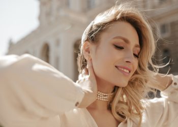 Cheerful blonde curly woman in pearl necklace and white elegant blouse smiles sincerely, looks down and poses outside.