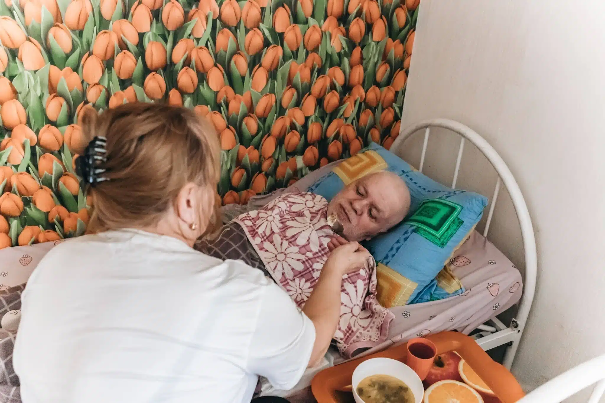 Сиделица ухаживает за пожилой женщиной в постели, помогая ей с едой и комфортом в уютной комнате с цветочным декором