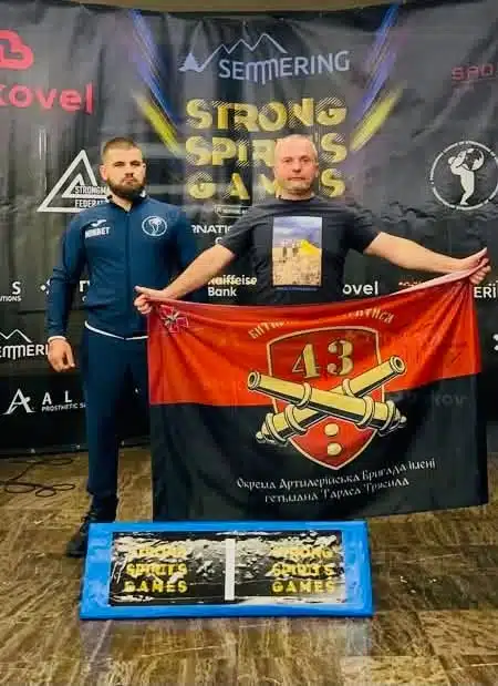 Два учасники тримають червоний військовий прапор 43ОАбр Тараса Трясила перед банером Strong Spirit's Games Сильні Переяслава