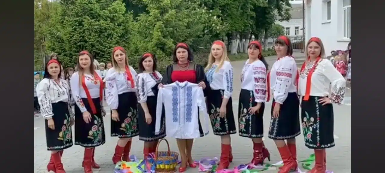 Дев'ять жінок у традиційних українських костюмах стоять в ряд для групового фото на подвір'ї