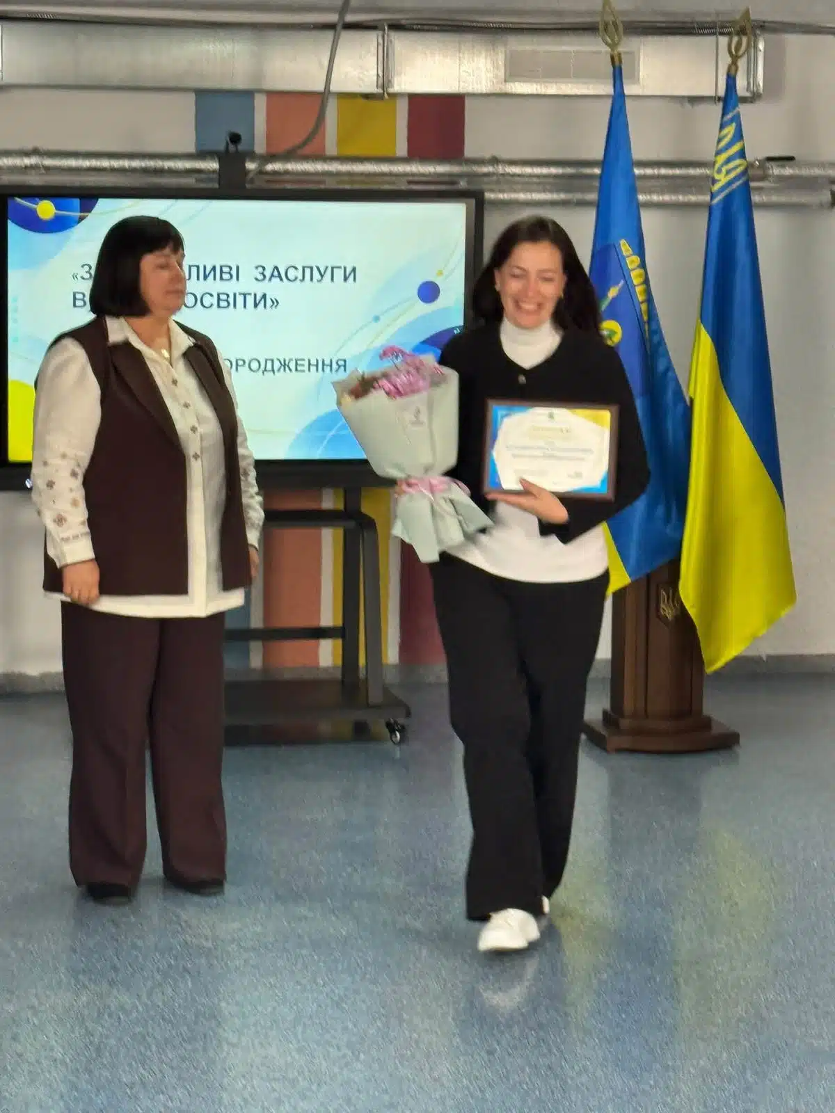 Жінка тримає сертифікат і букет від іншої жінки біля українського прапора