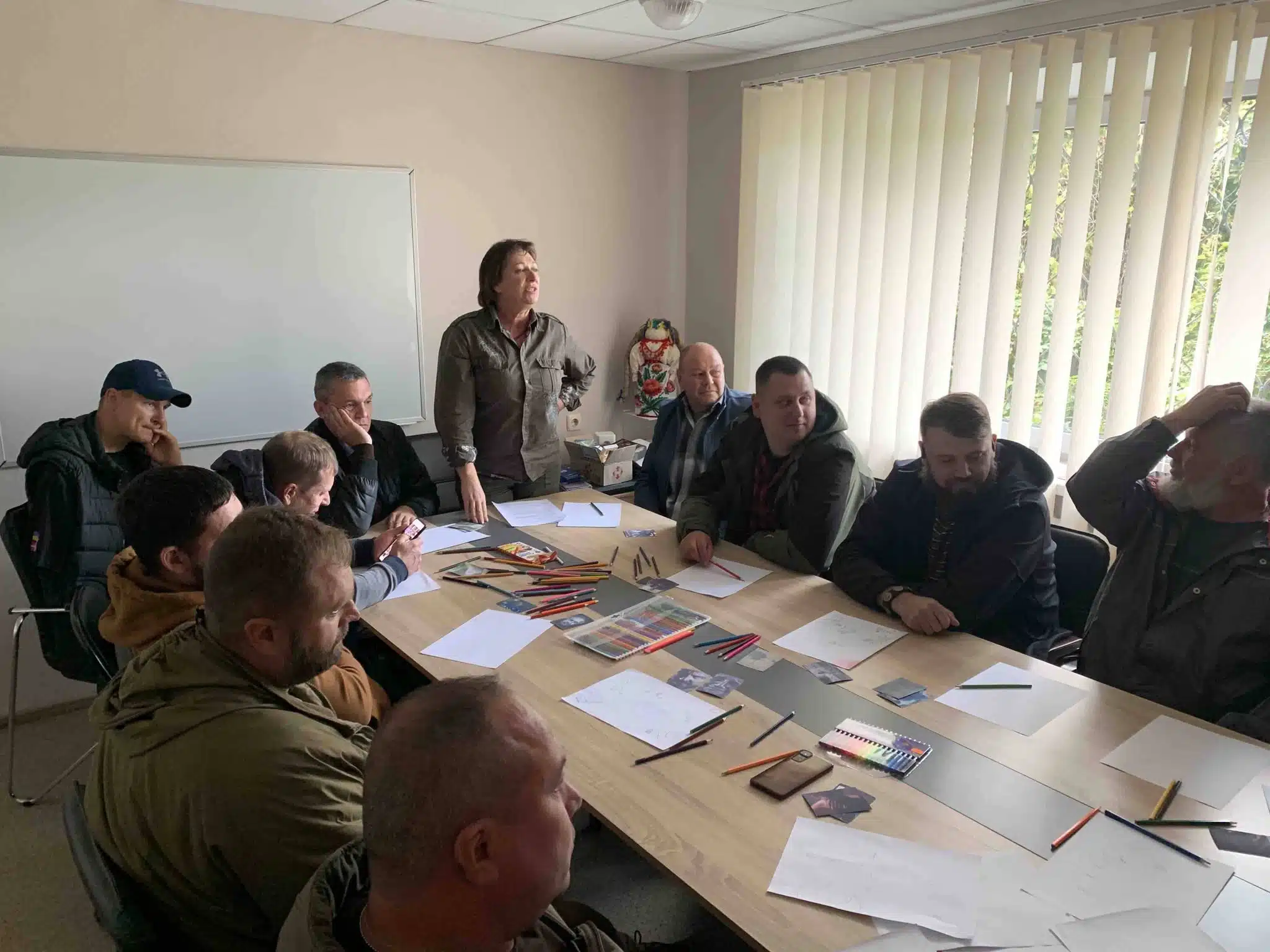 Група ветеранів та організаторів зустрічі стоять навколо столу з малюнками та творчими матеріалами в приміщенні з великими