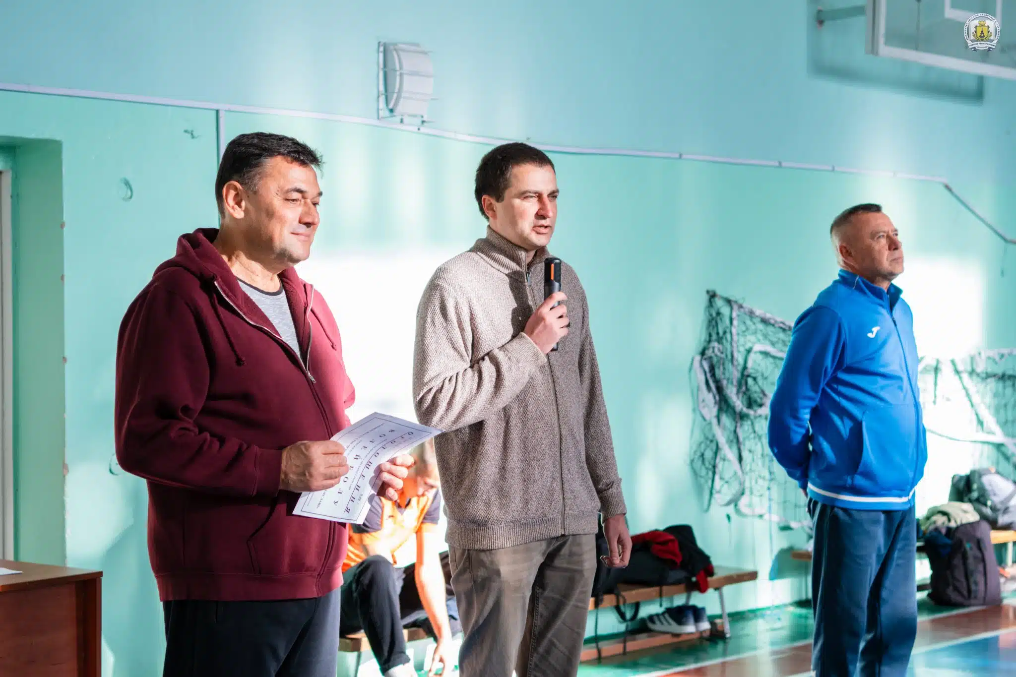 Три чоловіки у спортивному залі, один з мікрофоном звертається до учасників змагань