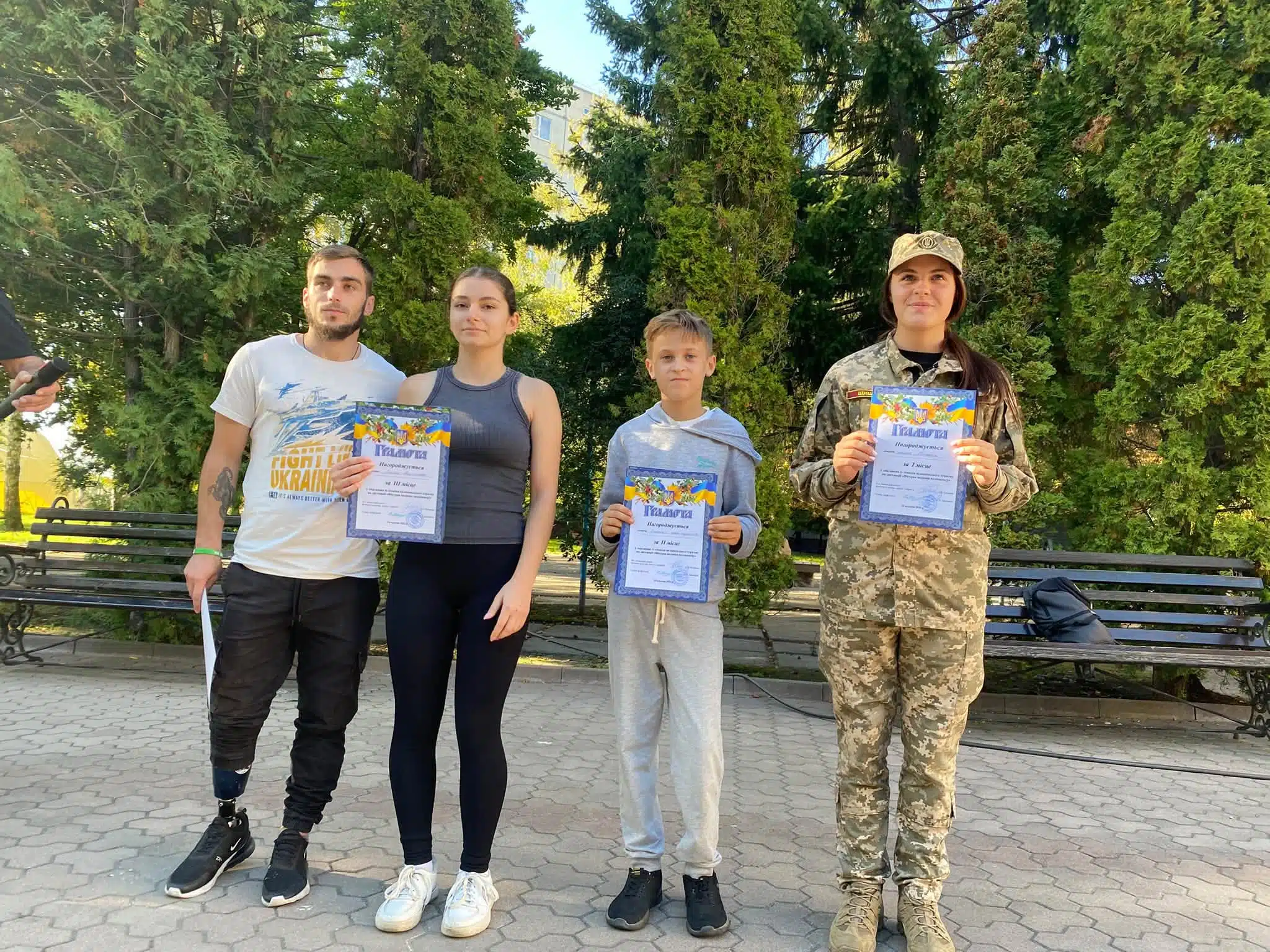 Четверо переможців зі сертифікатами стоять разом на бетонній доріжці на тлі дерев